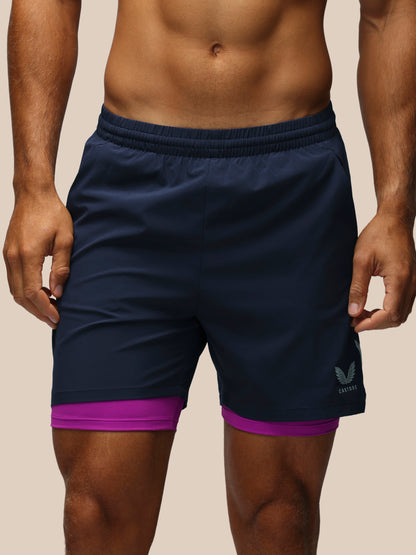 A close-up of a man wearing navy blue athletic shorts with a purple inner layer peeking out at the hem, featuring a small white logo on the lower left leg.
