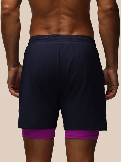 Rear view of a man wearing dark navy athletic shorts with a purple compression liner peeking out at the hem, against a solid beige background.