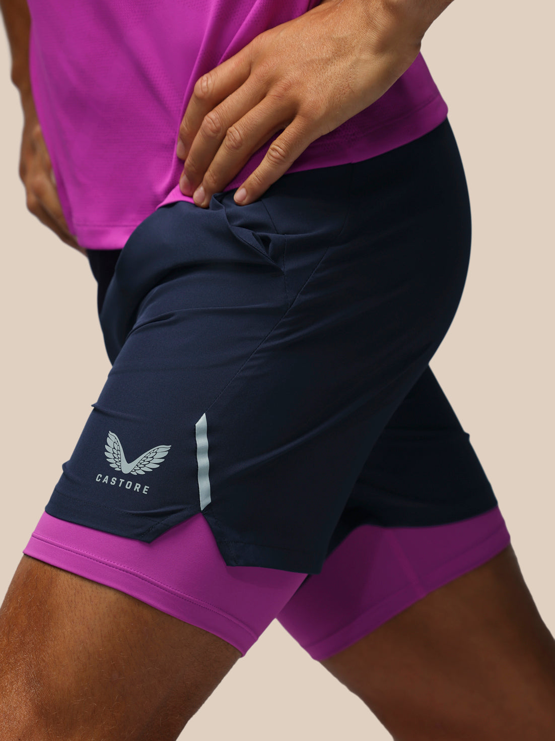 Close-up of a person wearing navy athletic shorts with a bright purple inner layer and a matching purple top, shown against a plain beige background. The shorts have a white wing logo and the brand name CASTORE visible on the lower left leg.