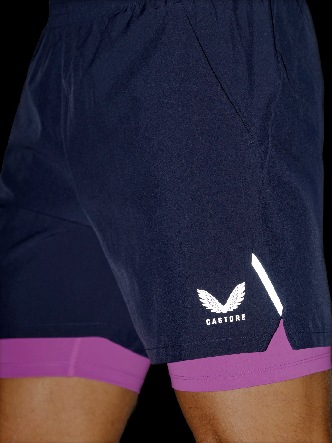 Close-up of a person wearing dark navy athletic shorts with a purple inner layer visible at the hem and a white wing logo with the word CASTORE on the lower left leg.