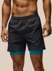 A man wearing dark charcoal gray athletic shorts with a teal inner layer visible at the hem, shown from the chest down against a plain beige background.