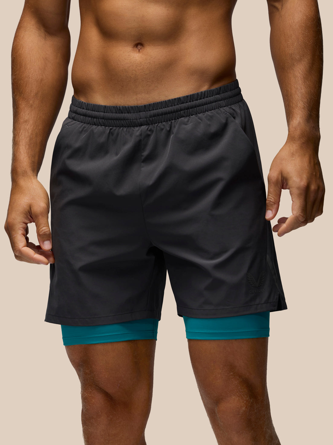 A man wearing dark charcoal gray athletic shorts with a teal inner layer visible at the hem, shown from the chest down against a plain beige background.