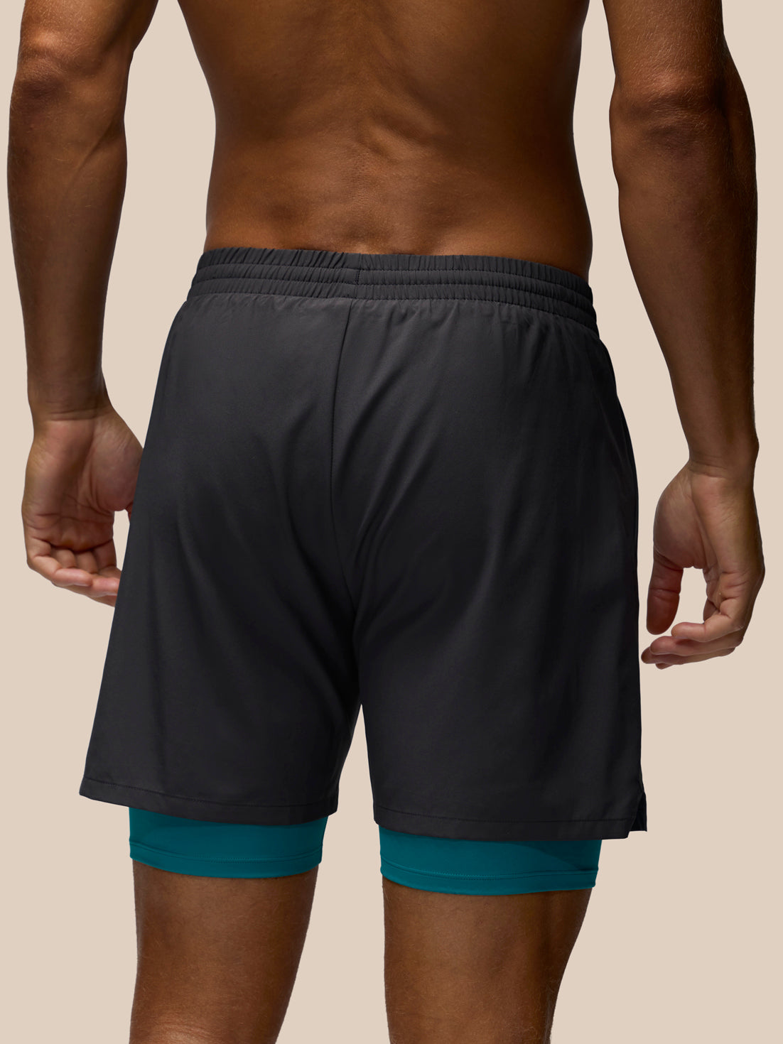 Rear view of a man wearing dark charcoal gray athletic shorts with a teal inner layer visible at the hem, against a plain beige studio background.