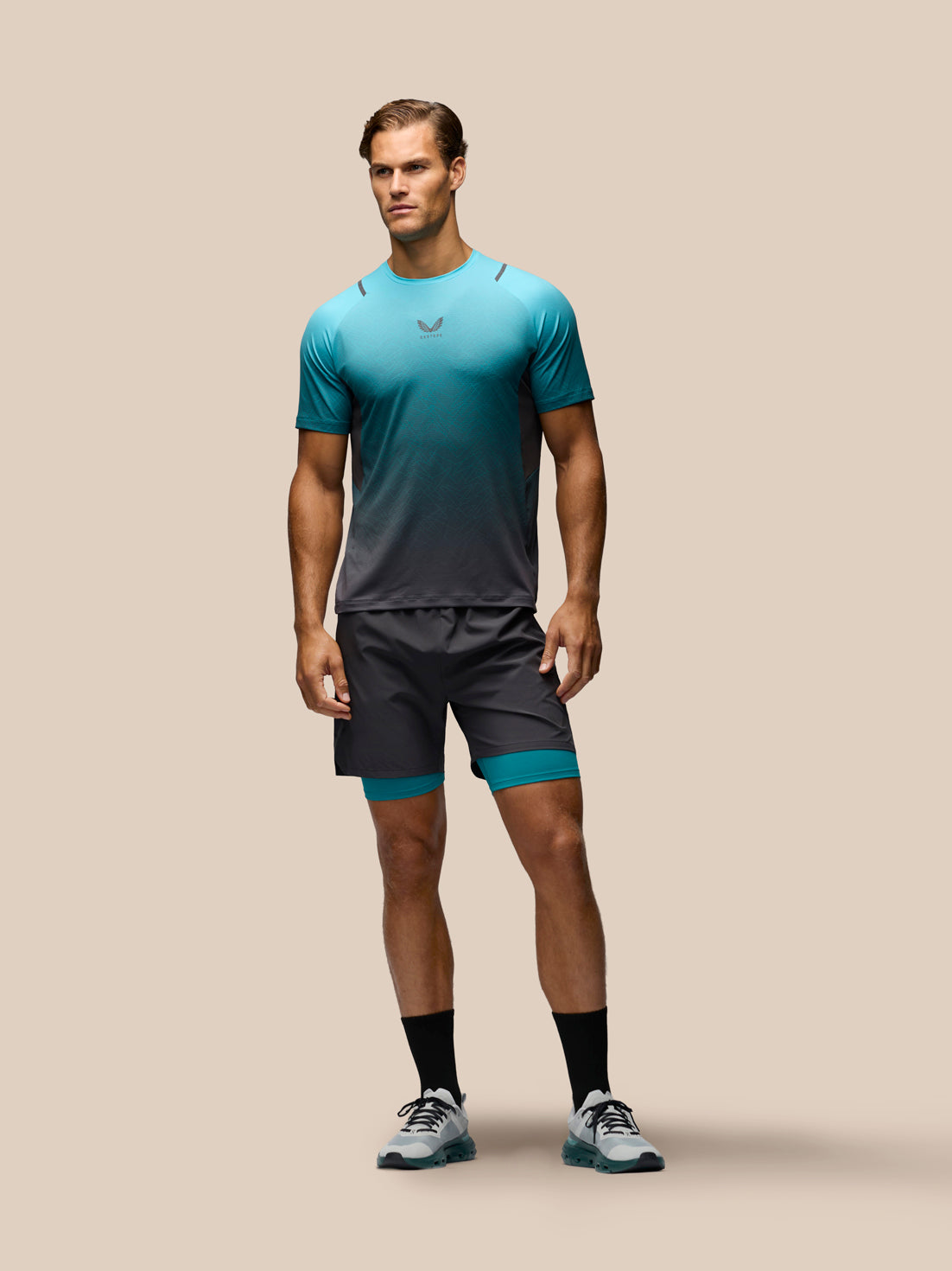 A male model stands against a solid beige background wearing a teal-to-dark-gray gradient athletic t-shirt, matching dark gray athletic shorts with teal inner leg bands, black crew socks, and gray running shoes.