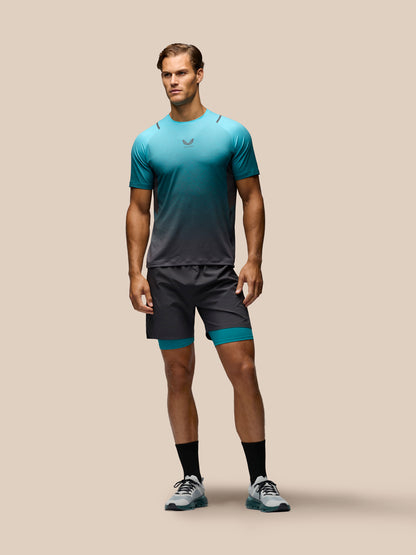 A male model stands against a solid beige background wearing a teal-to-dark-gray gradient athletic t-shirt, matching dark gray athletic shorts with teal inner leg bands, black crew socks, and gray running shoes.