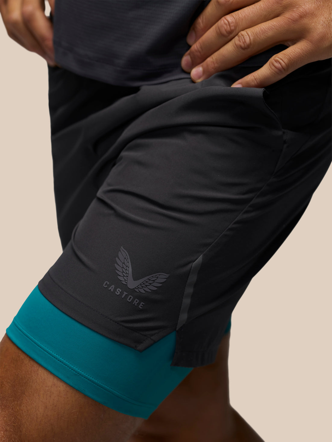 Close-up of a person wearing dark charcoal athletic shorts with a teal inner layer visible at the hem, plus a dark top. The shorts show a subtle wing logo and the word CASTORE near the lower left leg.