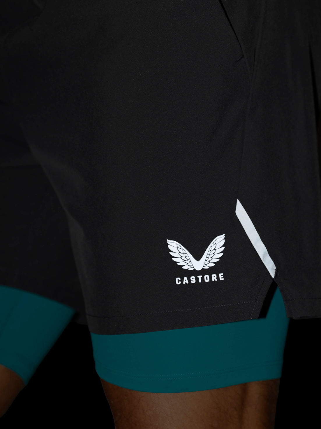 Close-up of black athletic shorts with a white Castore wing logo and a white reflective stripe, worn over teal compression leggings.