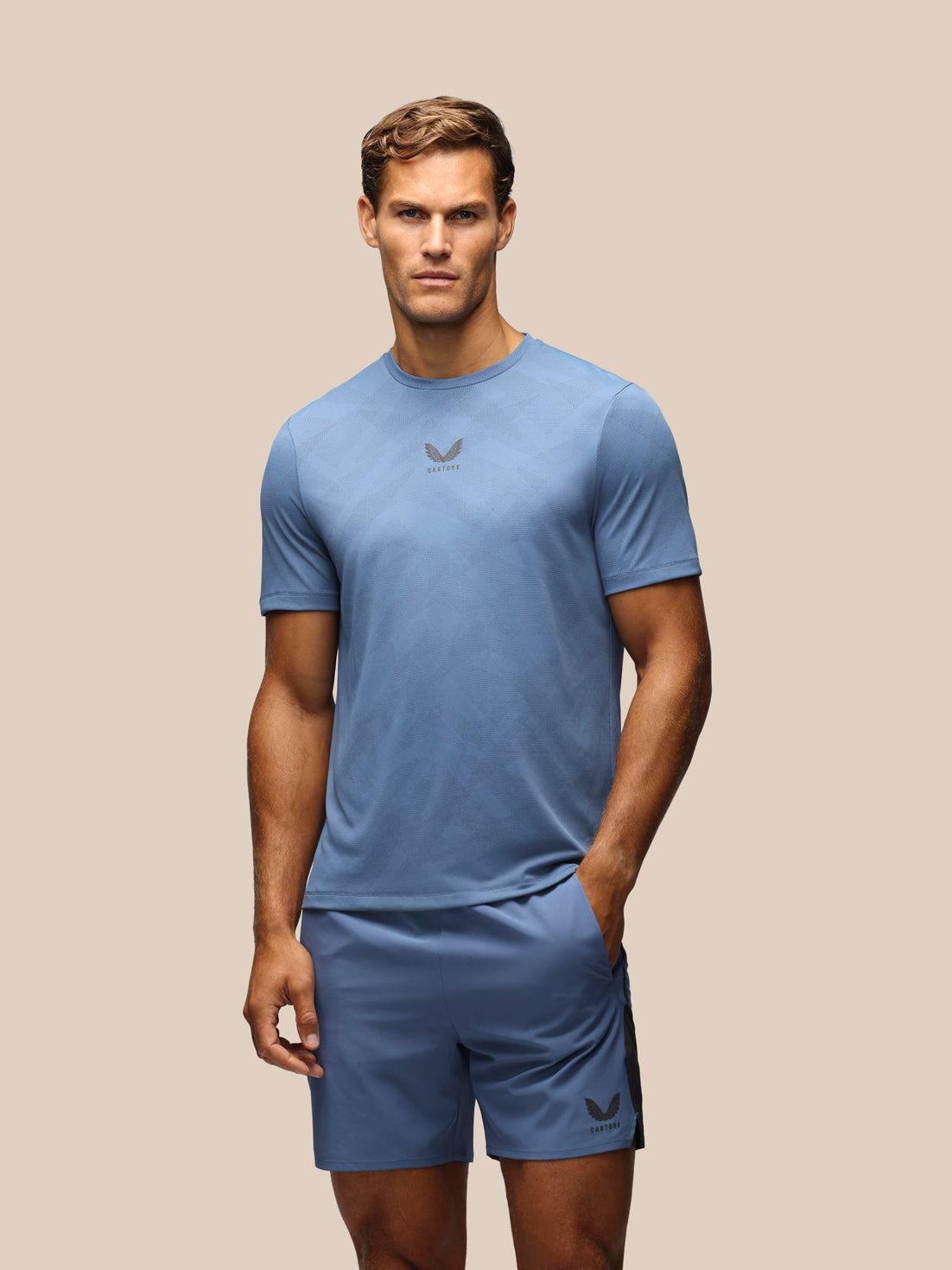 A man modeling a light blue athletic t-shirt and matching shorts with a dark gray Castore logo, set against a solid beige background.