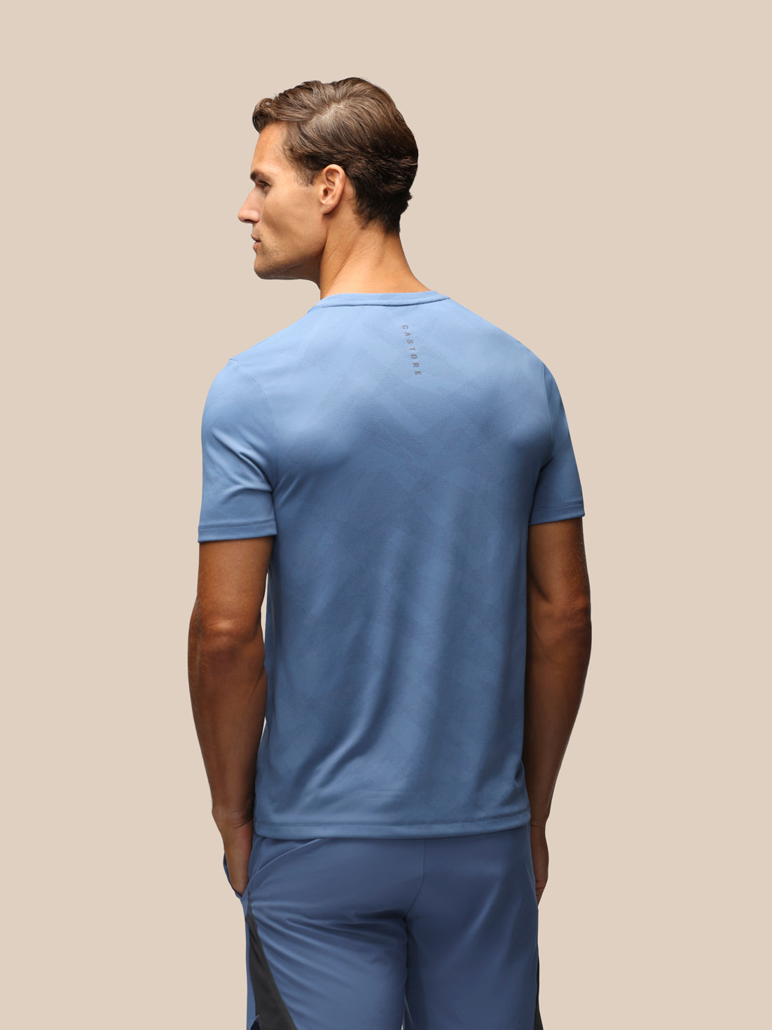A man shown from the back wearing a light blue short-sleeve athletic t-shirt and matching blue athletic pants, set against a solid beige background.