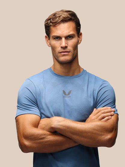 A muscular man with short brown hair wearing a light blue athletic t-shirt with a dark gray wing logo on the chest, standing with arms crossed against a solid beige background.