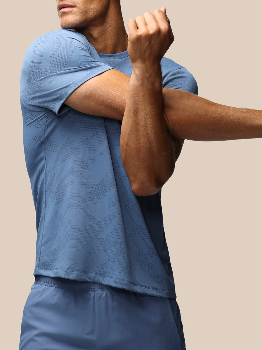 A man wearing a slate blue short-sleeve athletic t-shirt and matching blue shorts, shown from the chest down in a studio setting with a solid beige background.