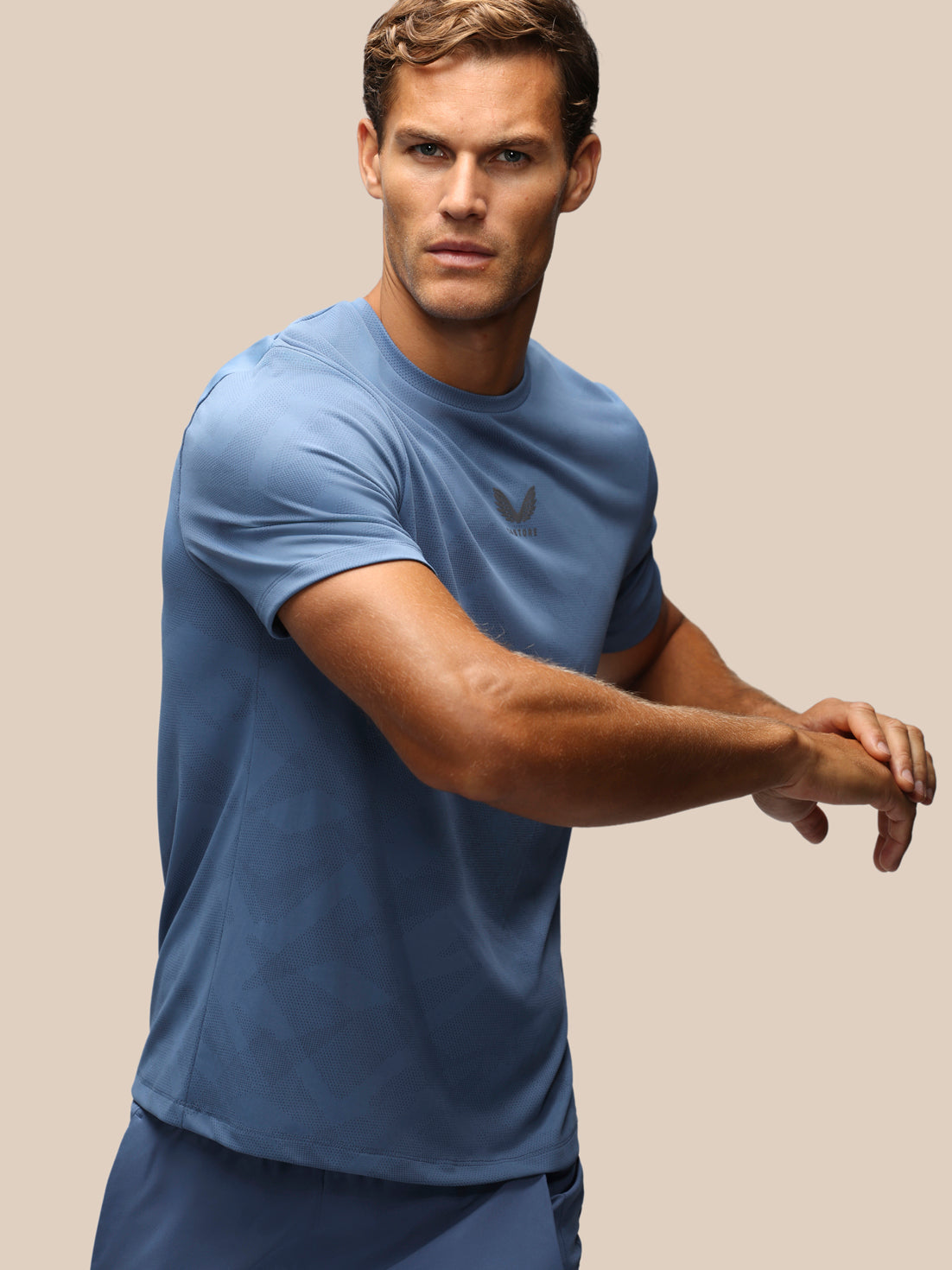A man wearing a blue athletic short-sleeve t-shirt with a small dark logo on the left chest, posed against a solid beige background.