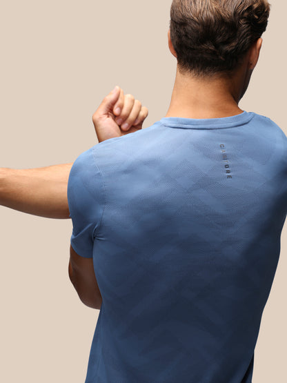 A man shown from behind wearing a blue short-sleeve t-shirt, highlighting the back design with a small vertical logo and a subtle diamond pattern.