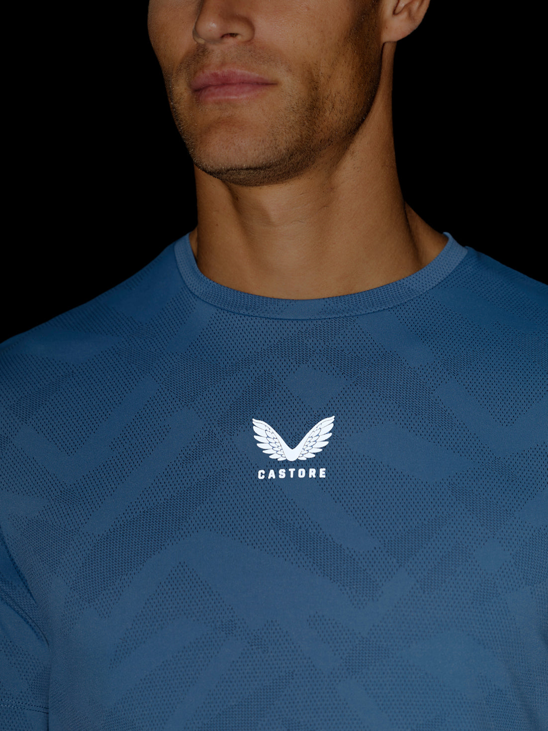 Close-up of a man wearing a blue textured athletic t-shirt with a white winged logo and the brand name 'CASTORE' printed on the chest, set against a solid black background.