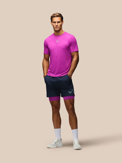 A male model stands against a solid beige background wearing a bright magenta athletic t-shirt, navy blue athletic shorts with a matching magenta inner layer, white socks, and white sneakers.