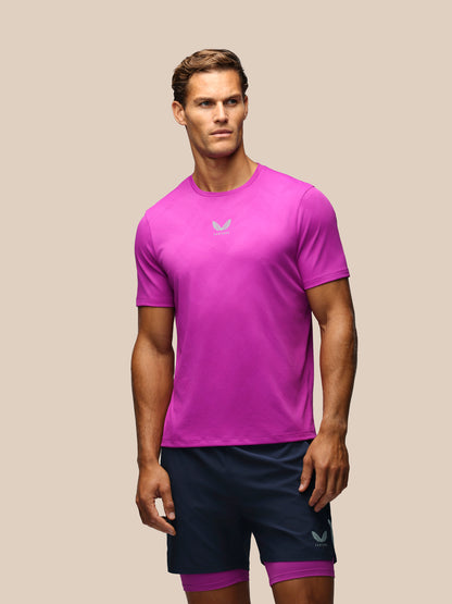 A male model wearing a bright magenta athletic t-shirt and navy blue athletic shorts with a purple inner layer, standing against a solid beige background.