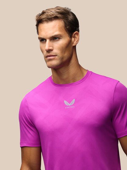 A man wearing a vibrant magenta athletic t-shirt with a small silver wing logo and the brand name 'Castore' printed on the chest, set against a solid beige background.