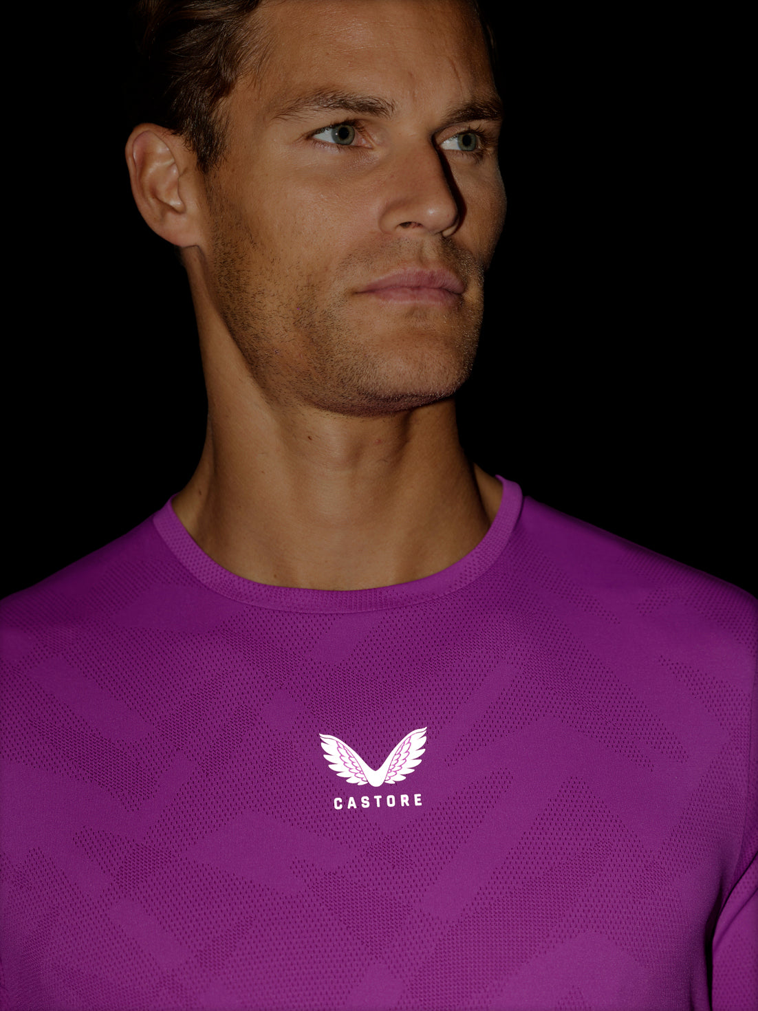 A close-up of a man wearing a purple Castore athletic t-shirt with a white wing logo and brand name on the chest, set against a solid black background.