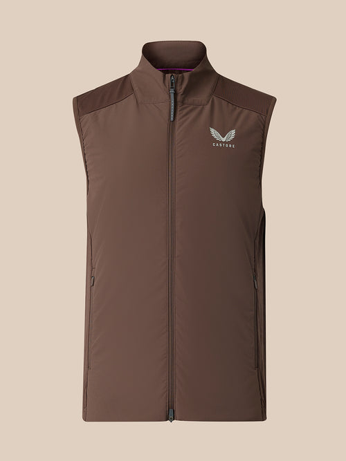 A sleeveless zip-front vest in dark brown with a stand collar and a small white wing logo on the left chest, shown against a plain beige background.