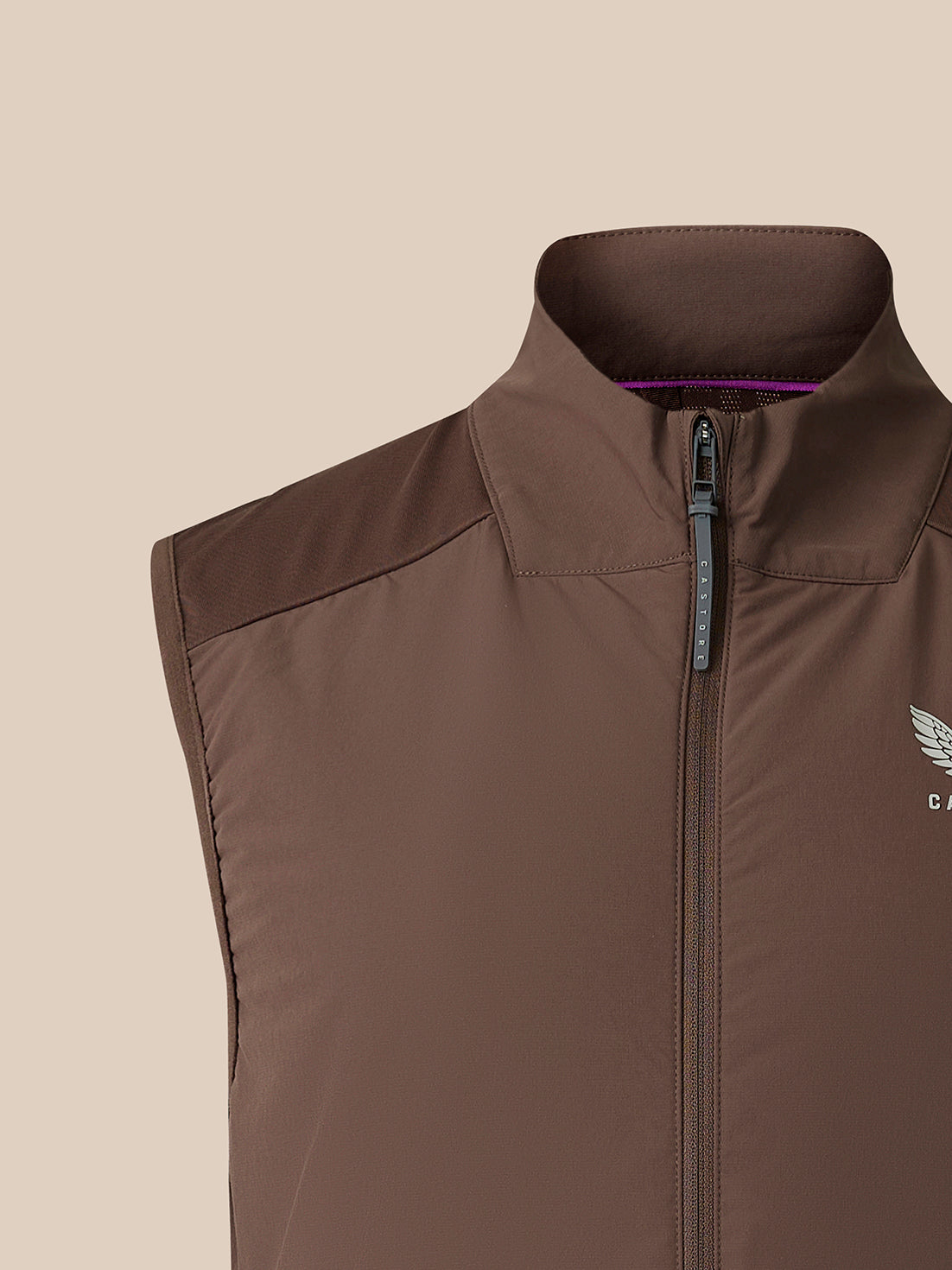 A close-up of a brown sleeveless zip-up vest with a high collar, featuring a small white wing logo on the left chest, set against a solid beige background.