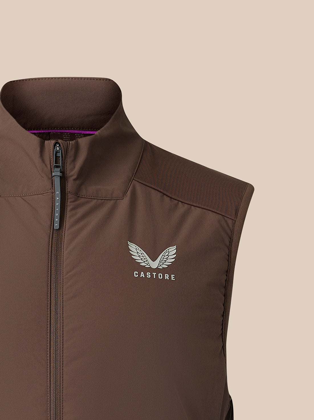 A close-up of a brown sleeveless zip-front vest with a stand collar, featuring a silver Castore wing logo on the chest, set against a solid beige background.