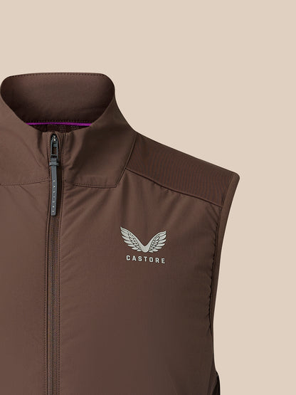 A close-up of a brown sleeveless zip-front vest with a stand collar, featuring a silver Castore wing logo on the chest, set against a solid beige background.