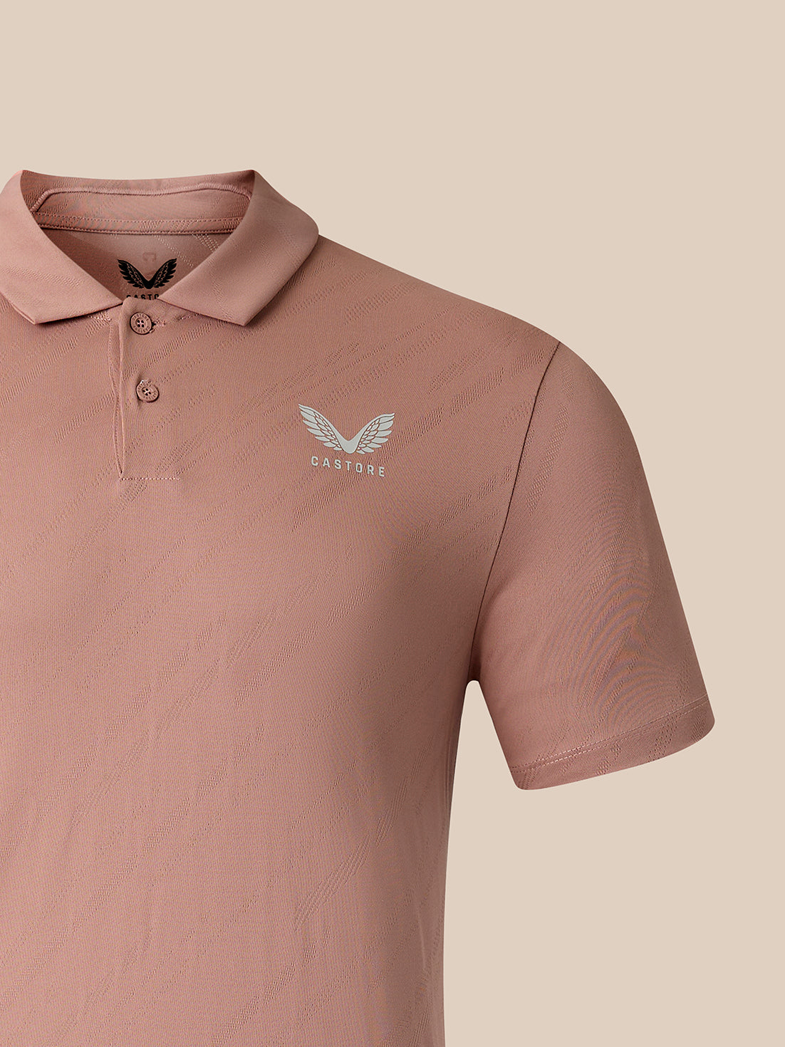 A close-up of a dusty rose pink polo shirt featuring a white embroidered winged logo with the word 'CASTORE' on the left chest, set against a solid beige background.