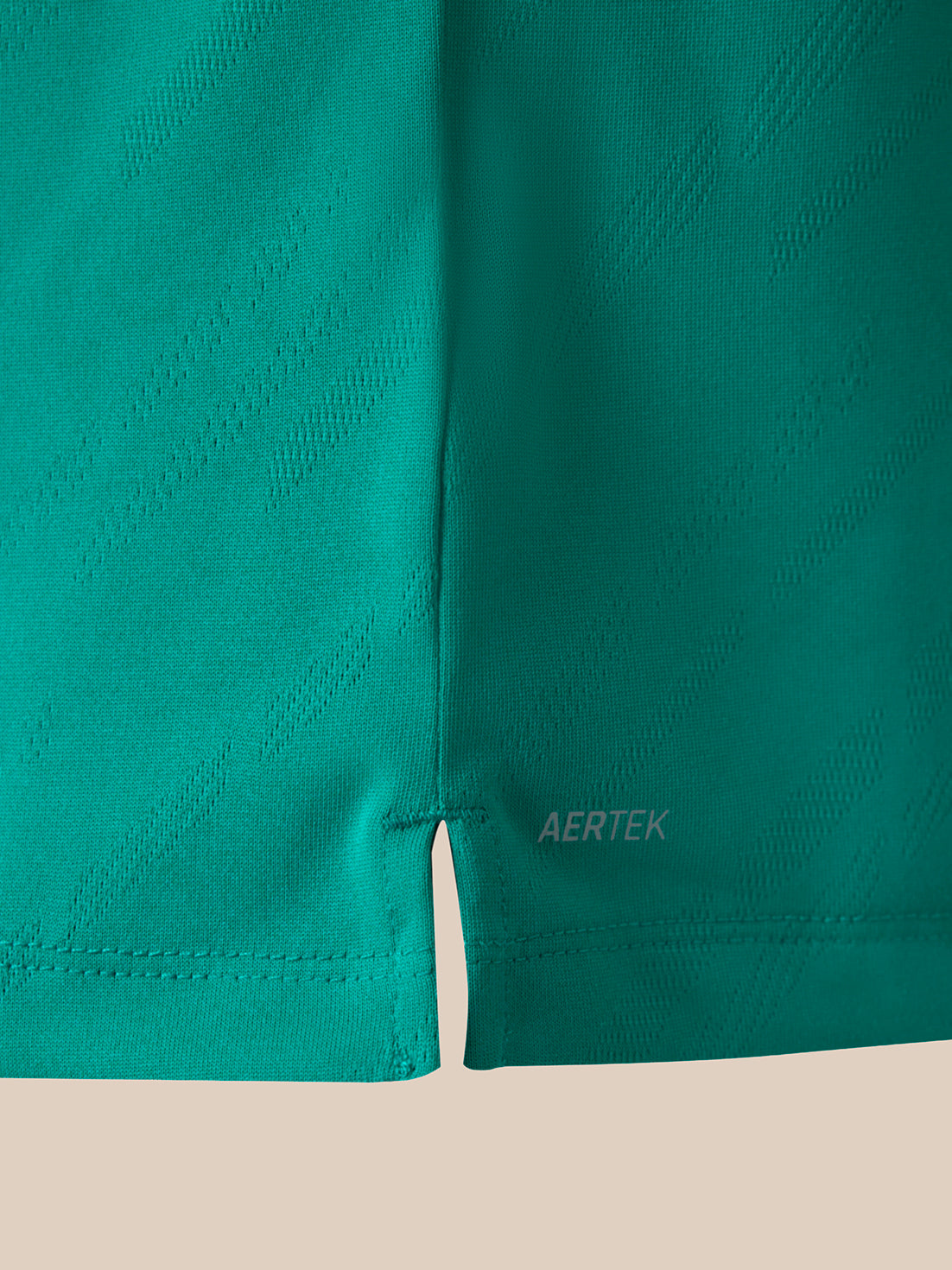 Close-up of teal athletic shirt showing the hem, stitched edge, and a small AERTEK logo near the lower right leg, set against a light beige background.