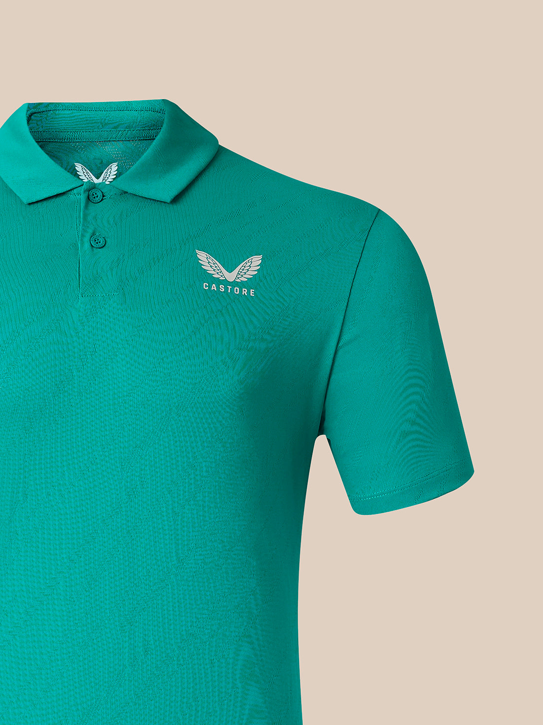A close-up of a teal short-sleeve polo shirt with a subtle textured pattern, featuring a white embroidered winged logo and the brand name 'CASTORE' on the left chest, set against a solid beige background.