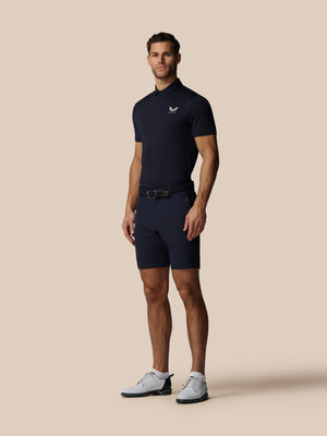 Man wearing a navy blue polo shirt and shorts with a belt on a beige background