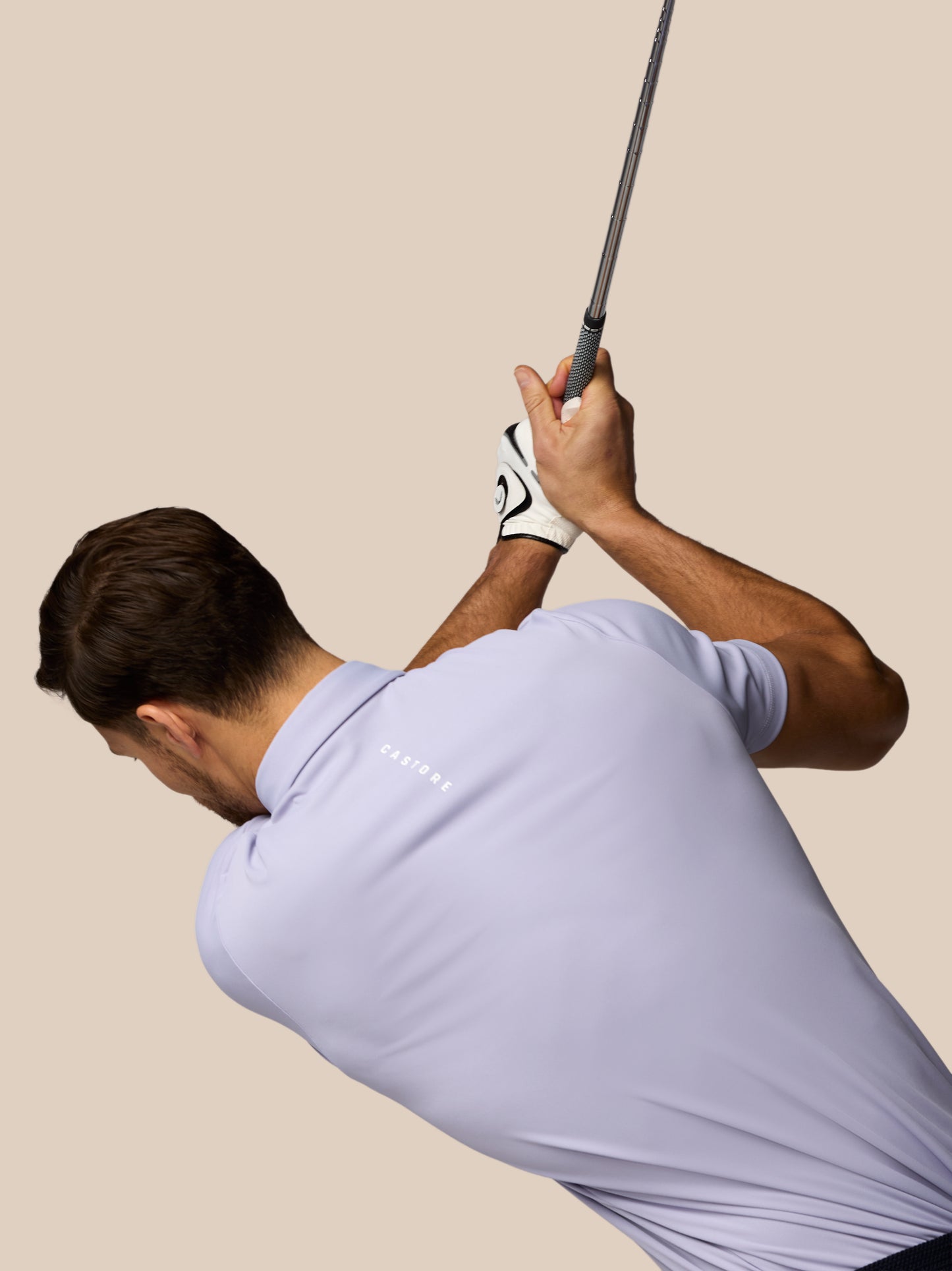 Man in a light purple shirt swinging a golf club against a beige background