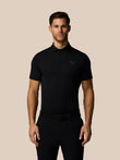 Male model wearing black Men’s Golf Essential polo showing chest Castore logo and moisture-wicking AerTek fabric against beige backdrop