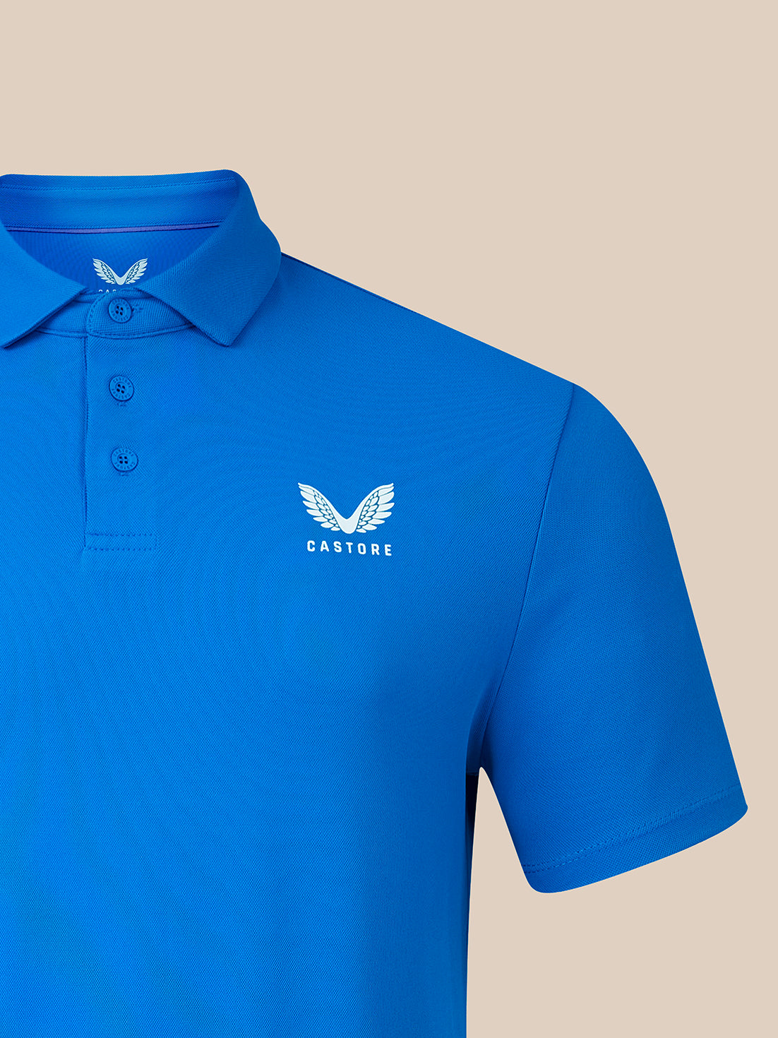 A bright blue short-sleeve polo shirt with a three-button placket and a white embroidered winged logo with the word 'CASTORE' on the left chest, shown against a solid beige background.