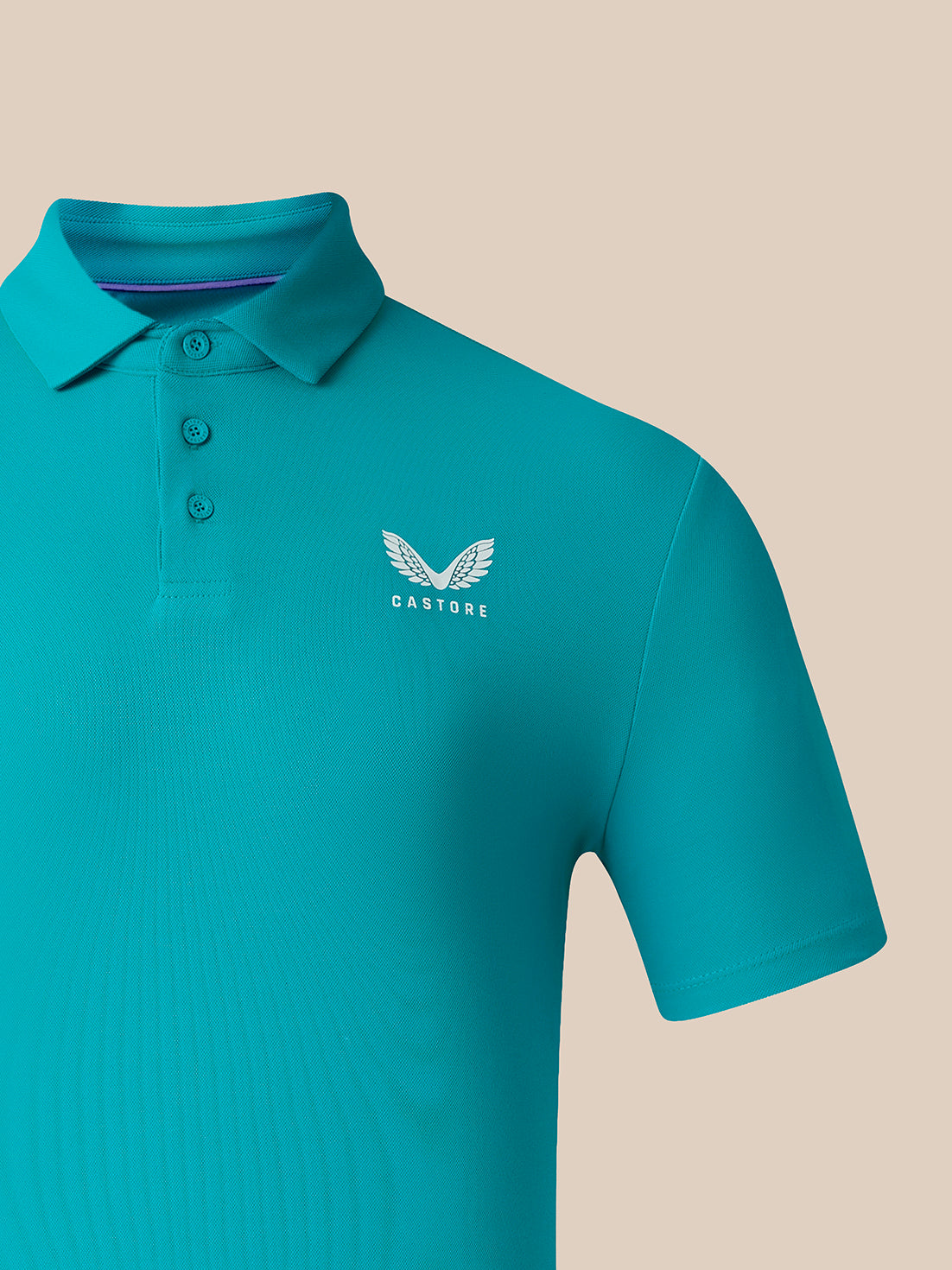 A close-up of a teal short-sleeve polo shirt with a three-button placket and a white embroidered winged logo with the brand name 'CASTORE' on the left chest, set against a solid beige background.