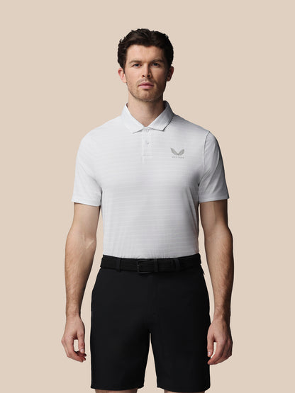 A man modeling a white short-sleeve polo shirt with a subtle horizontal stripe pattern and a small gray wing logo on the left chest, paired with black shorts, against a solid beige background.