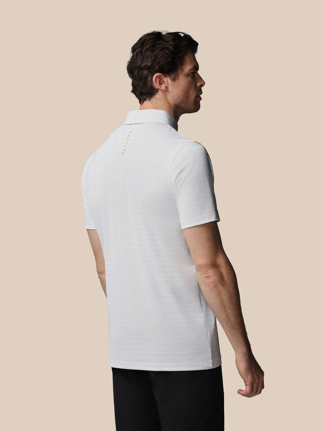 A man shown from the back wearing a white short-sleeve polo shirt and black pants, standing against a solid beige background.