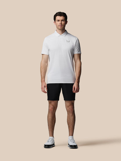 A man modeling a white short-sleeve polo shirt with a small embroidered logo on the left chest, paired with black athletic shorts and white sneakers, against a solid beige background.