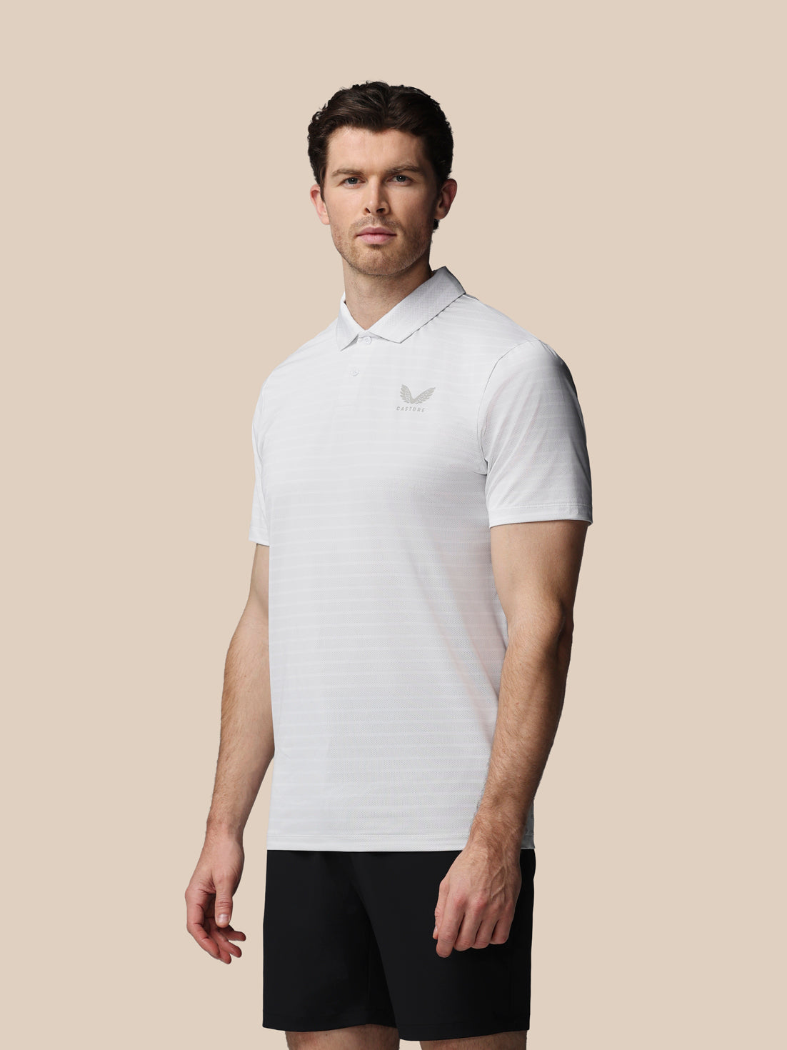 A man wearing a white short-sleeve polo shirt with a small gray embroidered logo on the left chest, paired with black shorts, set against a solid beige background.