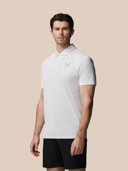 A man wearing a white short-sleeve polo shirt with a small gray embroidered logo on the left chest, paired with black shorts, set against a solid beige background.
