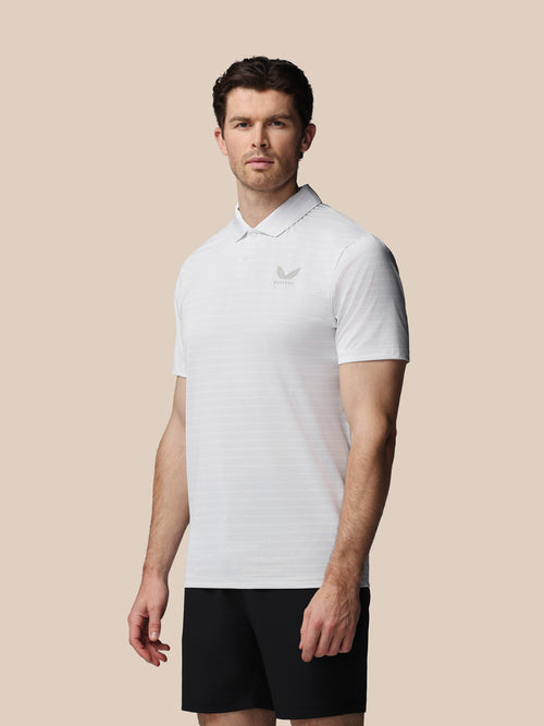 A man wearing a white short-sleeve polo shirt with a small gray embroidered logo on the left chest, paired with black shorts, set against a solid beige background.
