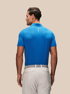 Back view of a man in a blue polo shirt with a vertical CASTSTORE logo on the upper back and light gray pants with a gray belt.