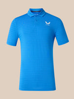 A bright blue short-sleeve polo shirt with a textured knit pattern, featuring a white wing logo and the brand name 'CASTORE' on the left chest, displayed against a solid beige background.