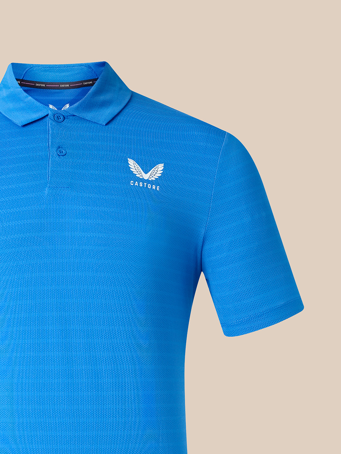 A bright blue short-sleeve polo shirt with a textured knit pattern, featuring a white winged logo and the brand name 'CASTORE' on the left chest, set against a solid beige background.