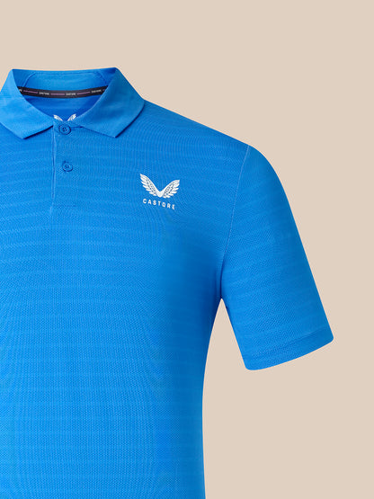 A bright blue short-sleeve polo shirt with a textured knit pattern, featuring a white winged logo and the brand name 'CASTORE' on the left chest, set against a solid beige background.