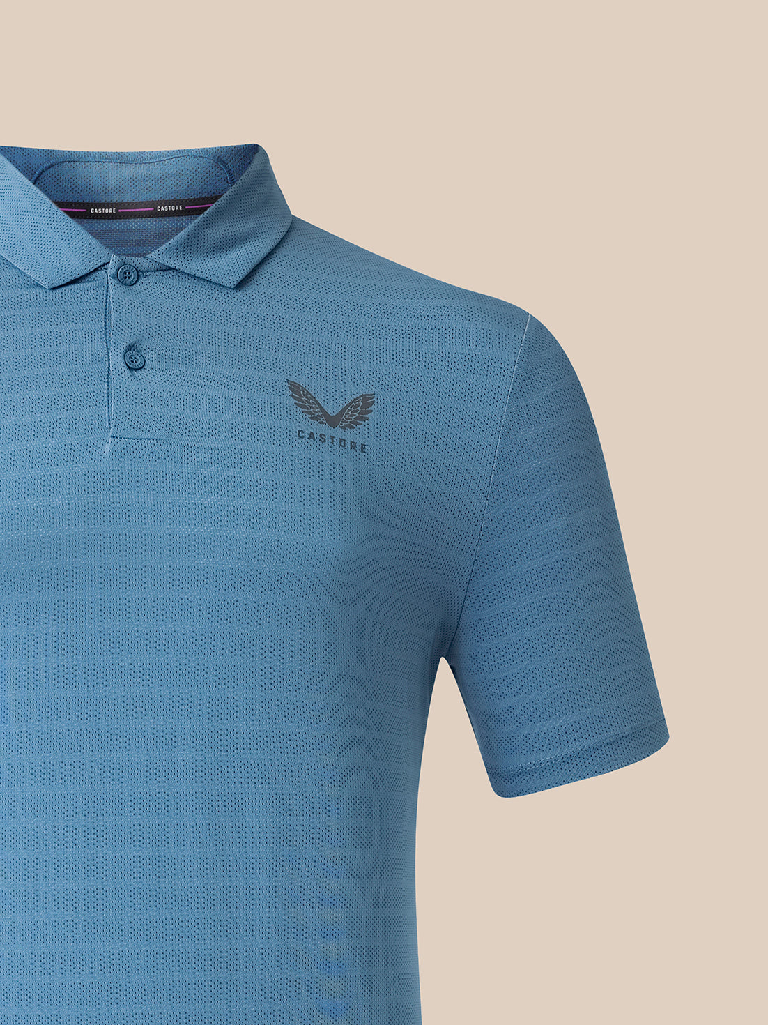 A close-up of a blue short-sleeve polo shirt with a subtle textured pattern, featuring a dark gray embroidered winged logo and the brand name 'CASTORE' on the left chest, set against a solid beige background.