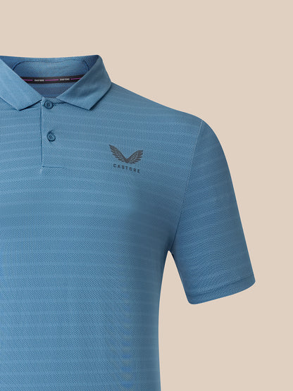 A close-up of a blue short-sleeve polo shirt with a subtle textured pattern, featuring a dark gray embroidered winged logo and the brand name 'CASTORE' on the left chest, set against a solid beige background.