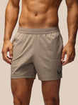 A close-up of a man wearing light taupe athletic shorts with a small black wing logo on the lower left leg, set against a solid beige background.
