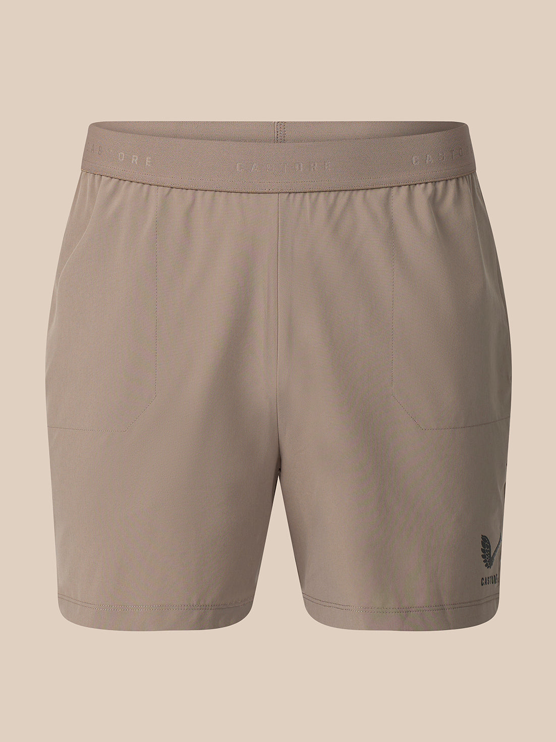 A pair of women's athletic shorts in a solid taupe color, featuring a wide elastic waistband with subtle branding and a small dark logo on the lower left leg, shown against a solid beige background.