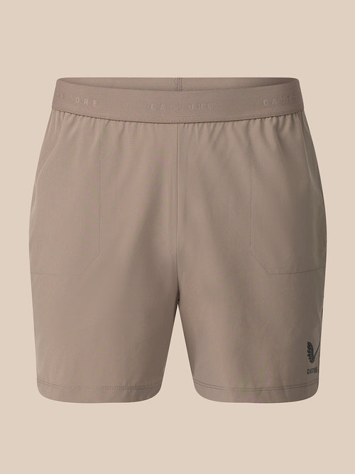 A pair of women's athletic shorts in a solid taupe color, featuring a wide elastic waistband with subtle branding and a small dark logo on the lower left leg, shown against a solid beige background.