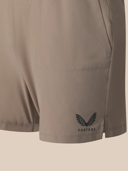 Close-up of a pair of light taupe athletic shorts showing a side pocket and a dark gray Castore wing logo on the lower left leg, set against a solid light beige background.